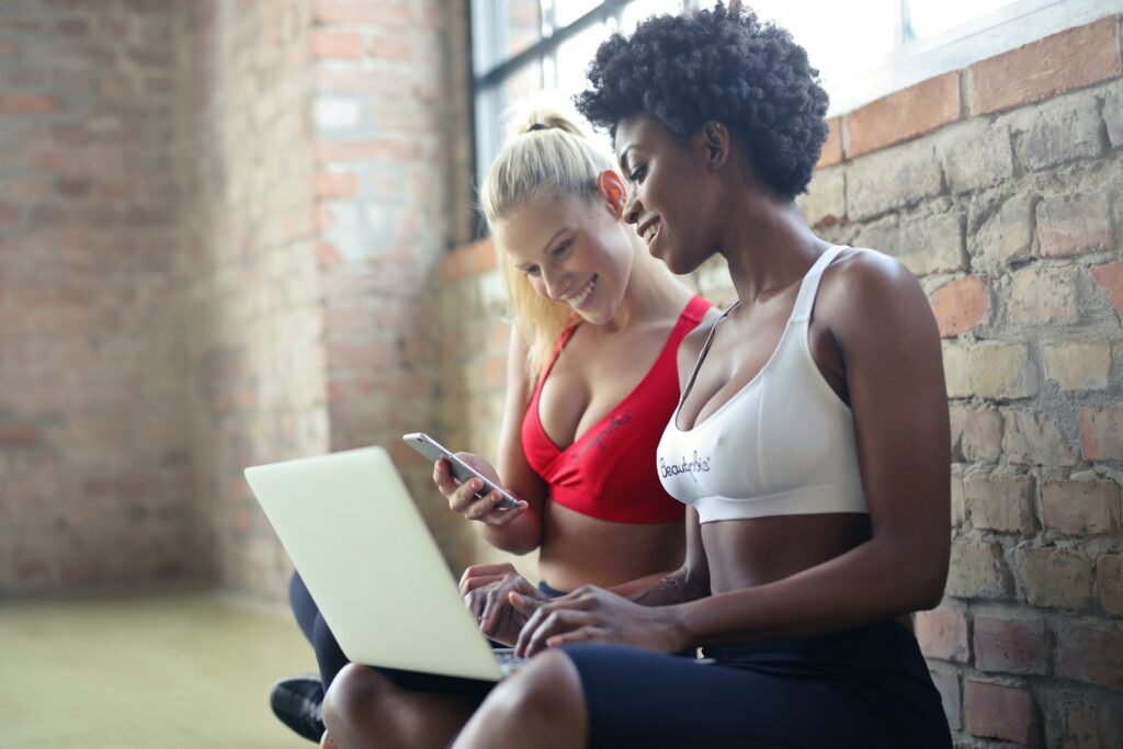 pexels-photo-863950-863950 Two women in sportswear enjoy leisure time, sharing a laptop and smartphone in a gym setting against a brick wall.